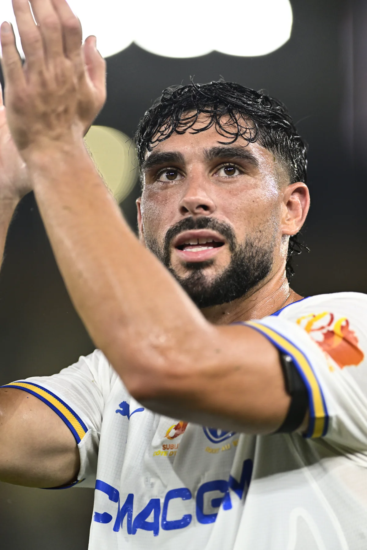 Joueur de l'OM en maillot blanc et bleu avec le numéro visible, levant le bras en célébration lors d'un match. Son visage exprime l'émotion et l'engagement sportif.