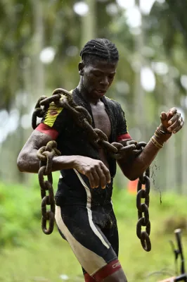 Jeune athlète musclé effectuant un entraînement intensif en tenant de lourdes chaînes métalliques sous la pluie, démontrant force et détermination dans un environnement naturel verdoyant.