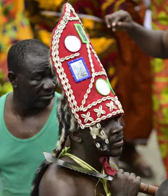 Portrait d'un homme portant une coiffe conique rouge décorée de coquillages blancs, de miroirs et de motifs traditionnels, avec des ornements corporels et une arme, lors d'une célébration culturelle