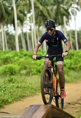 Cycliste portant un casque et des lunettes de soleil chevauchant un vélo orange et noir sur un sentier de terre en forêt tropicale avec des palmiers en arrière-plan