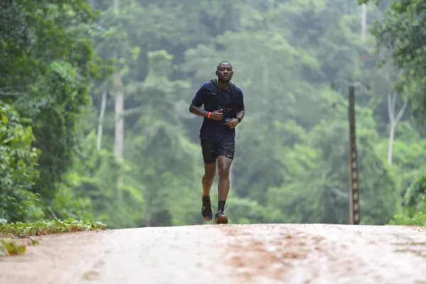 Un coureur de trail court sur un chemin de terre dans une forêt dense avec de grands arbres. Il porte un maillot noir et un short noir avec un brassard orange. Le paysage boisé s'étend en arrière-plan avec une atmosphère brumeuse.