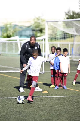 Un coach en tenue noire Nike supervise un jeune garçon qui tire un ballon de soccer sur un terrain synthétique, entouré d'autres enfants en tenue de sport bleu et rouge.