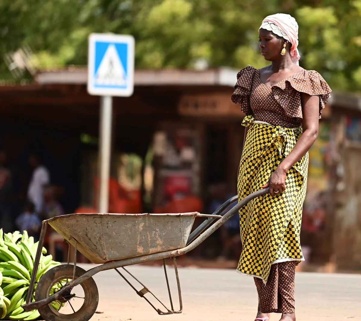 Une femme vêtue traditionnellement avec un foulard rose, une robe marron à pois et un pagne jaune à motifs tient une brouette sur une route devant des bâtiments flous et une signalisation routière