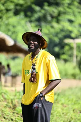 Homme souriant portant un maillot jaune Brésil, un chapeau de paille marron, une lanière avec pendentif et une montre. Arrière-plan avec végétation verte luxuriante et bâtiments flous.