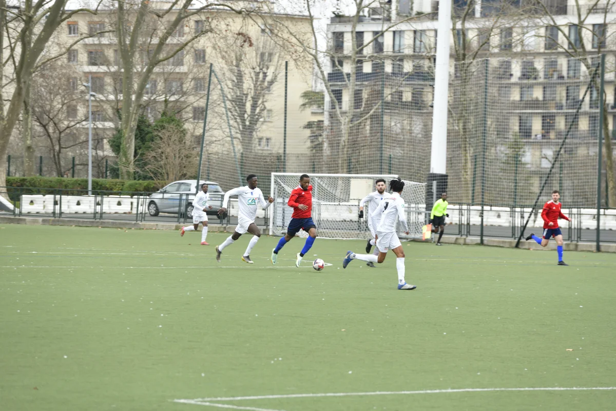 Match de football entre deux équipes, l'une en blanc et l'autre en rouge et bleu, jouant sur un terrain synthétique en ville avec des immeubles en arrière-plan