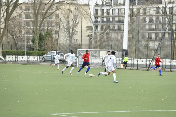 Match de football entre deux équipes, l'une en blanc et l'autre en rouge et bleu, jouant sur un terrain synthétique en ville avec des immeubles en arrière-plan