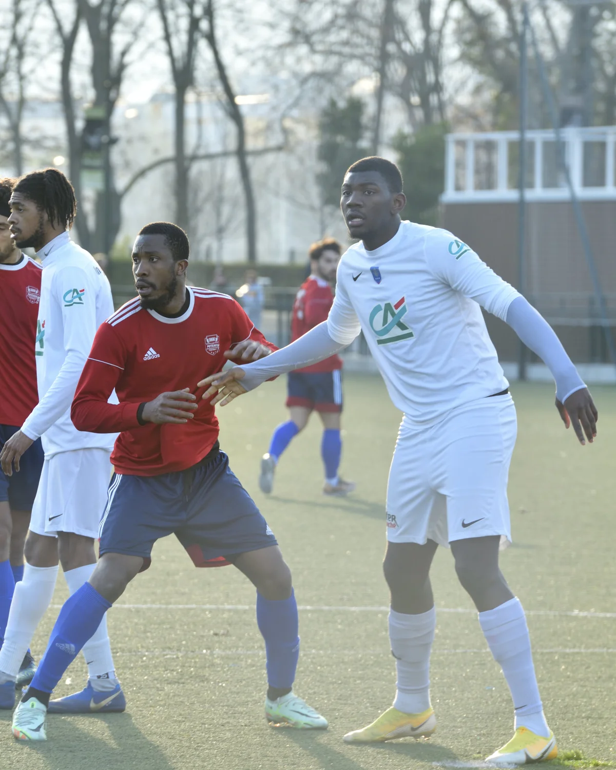 Joueurs de football pendant un match : un joueur en maillot rouge et short bleu fonce avec un joueur en maillot blanc et short bleu. Autres joueurs visibles en arrière-plan sur un terrain de sport.