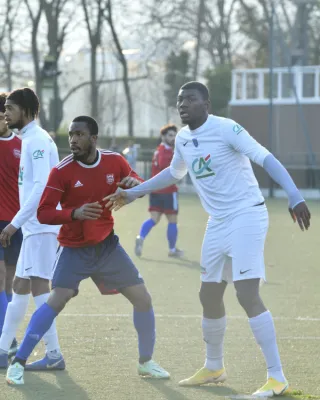 Joueurs de football pendant un match : un joueur en maillot rouge et short bleu fonce avec un joueur en maillot blanc et short bleu. Autres joueurs visibles en arrière-plan sur un terrain de sport.