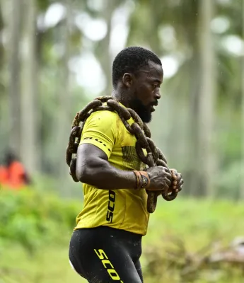 Athlète masculin portant une chaîne à dos et un gilet de trail jaune vif, courant en montée sous la pluie dans une forêt verdoyante avec des arbres en arrière-plan flou