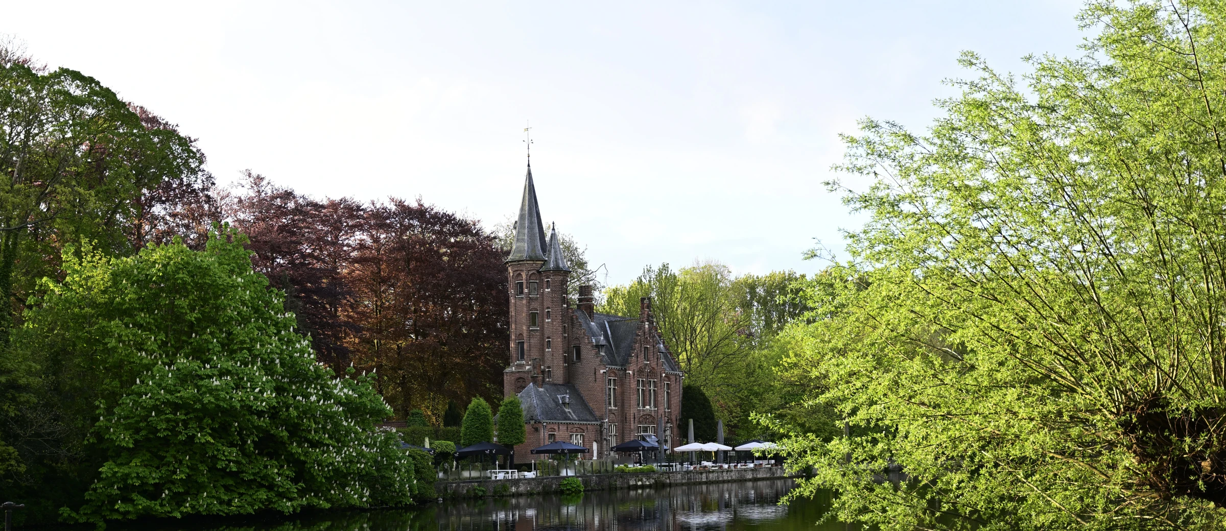 Un château de style gothique avec tours pointues en brique rouge situé au bord d'une rivière, entouré d'arbres feuillus verts et de terrasses blanches, photographié par temps clair
