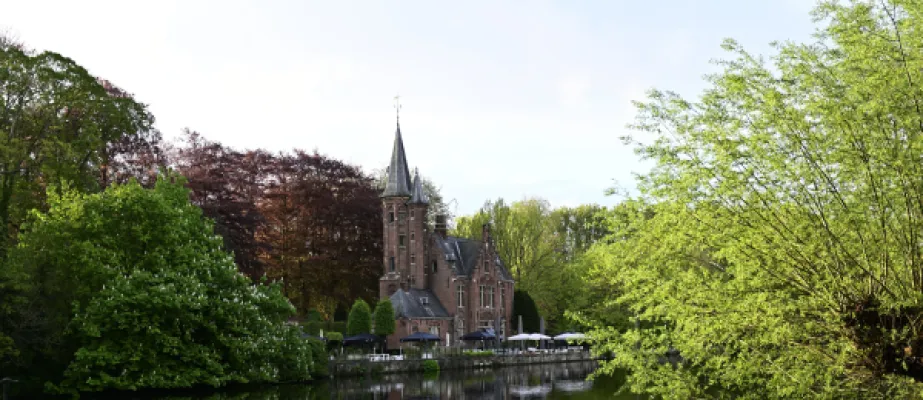 Un château de style gothique avec tours pointues en brique rouge situé au bord d'une rivière, entouré d'arbres feuillus verts et de terrasses blanches, photographié par temps clair