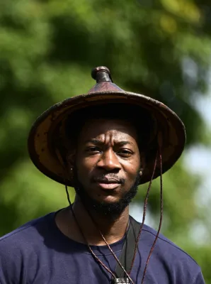 Homme portant un chapeau conique traditionnel et un collier, souriant face à la caméra avec un arrière-plan de verdure floue