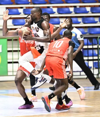 Joueur en maillot blanc et rouge effectue une passe lors d'un match de basketball en salle, défendu par un adversaire en rouge, devant des gradins bleus.