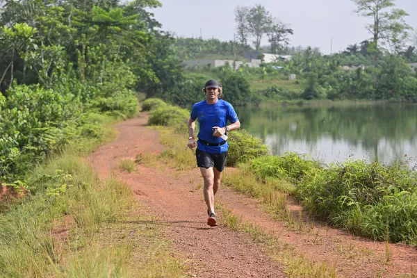 Coureur en chemise bleue et short noir pratiquant le trail running sur un chemin en terre rouge bordé de végétation luxuriante, avec un lac et des habitations en arrière-plan