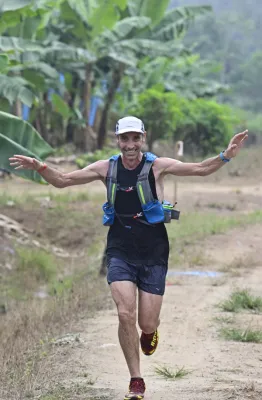 Athlète masculin souriant en train de courir sur un sentier de montagne, portant un gilet de trail bleu et blanc, bras écartés, avec des bananiers verts en arrière-plan