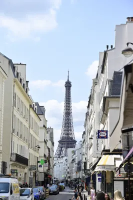 La Tour Eiffel vue du bout d'une rue parisienne bordée de bâtiments haussmanniens blancs et beige, avec commerces et piétons en bas de la rue par beau temps
