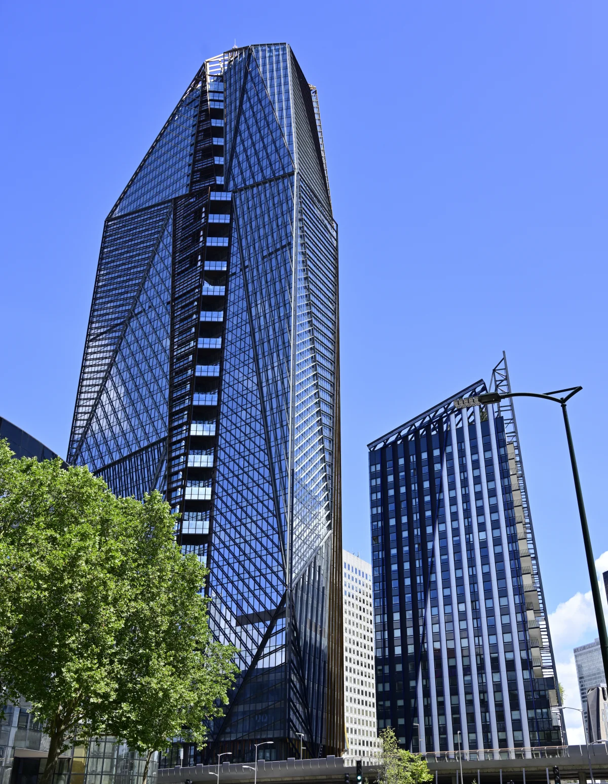 Immeuble de grande hauteur aux murs en verre bleu et noir avec design géométrique angulaire, entouré d'autres bâtiments modernes et d'arbres verts sous un ciel bleu clair