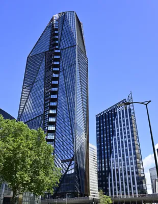 Immeuble de grande hauteur aux murs en verre bleu et noir avec design géométrique angulaire, entouré d'autres bâtiments modernes et d'arbres verts sous un ciel bleu clair